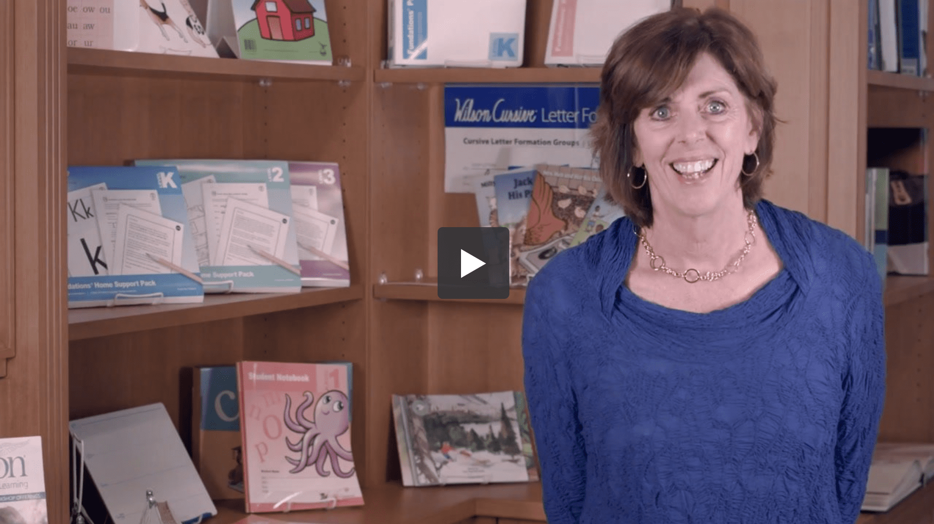 A woman with short brown hair wearing a blue blouse stands smiling in a room with shelves filled with books and educational materials. A video play button is visible at the center of the image, suggesting the frame is from a video.